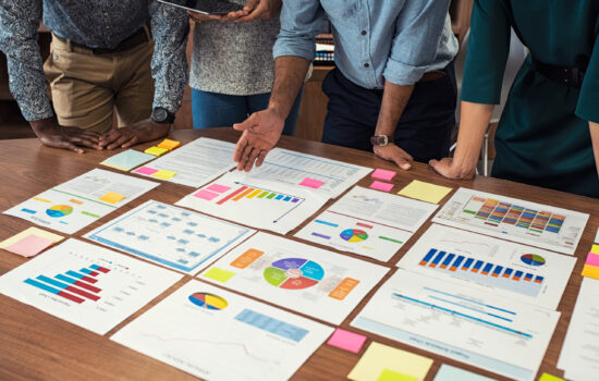 Financial and business documents on table with multiethnic hands working on it. Latin business manager with colleagues working on new startup project. Closeup business man and businesswoman hands understanding pie and bar graphs during meeting.